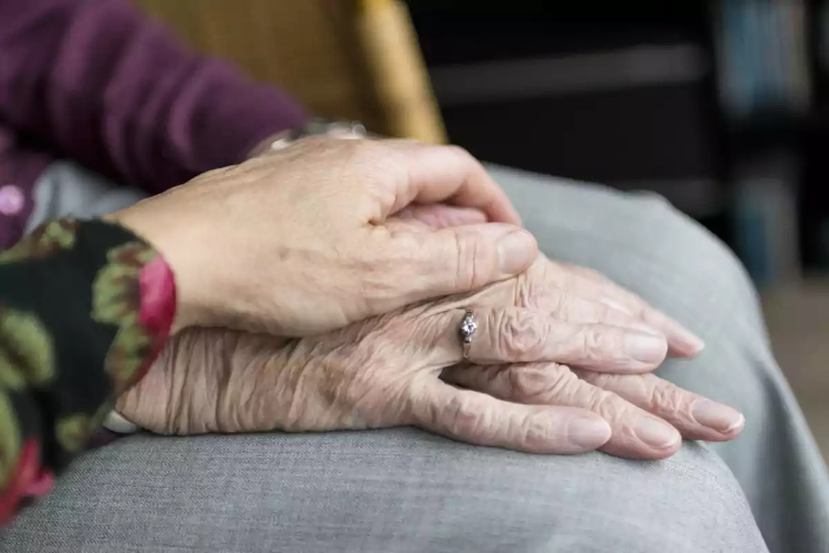 Mano de una persona mayor con un anillo siendo sostenida por otra mano en señal de apoyo y cariño