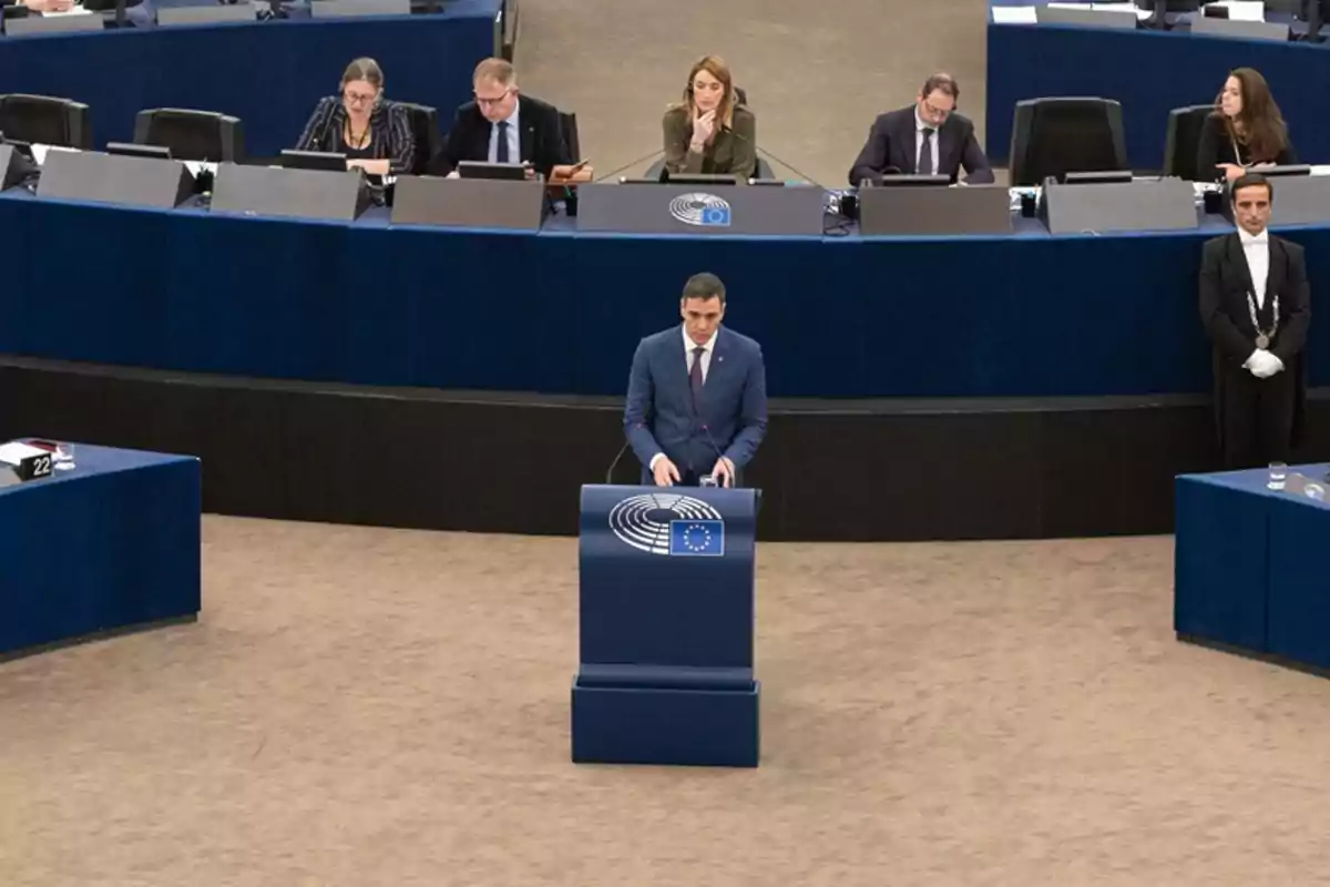 Un hombre de traje azul da un discurso en un podio azul con el logo del Parlamento Europeo mientras varias personas lo observan desde una mesa elevada al fondo Un hombre de traje azul da un discurso en un podio azul con el logo del Parlamento Europeo mientras varias personas lo observan desde una mesa elevada al fondo
