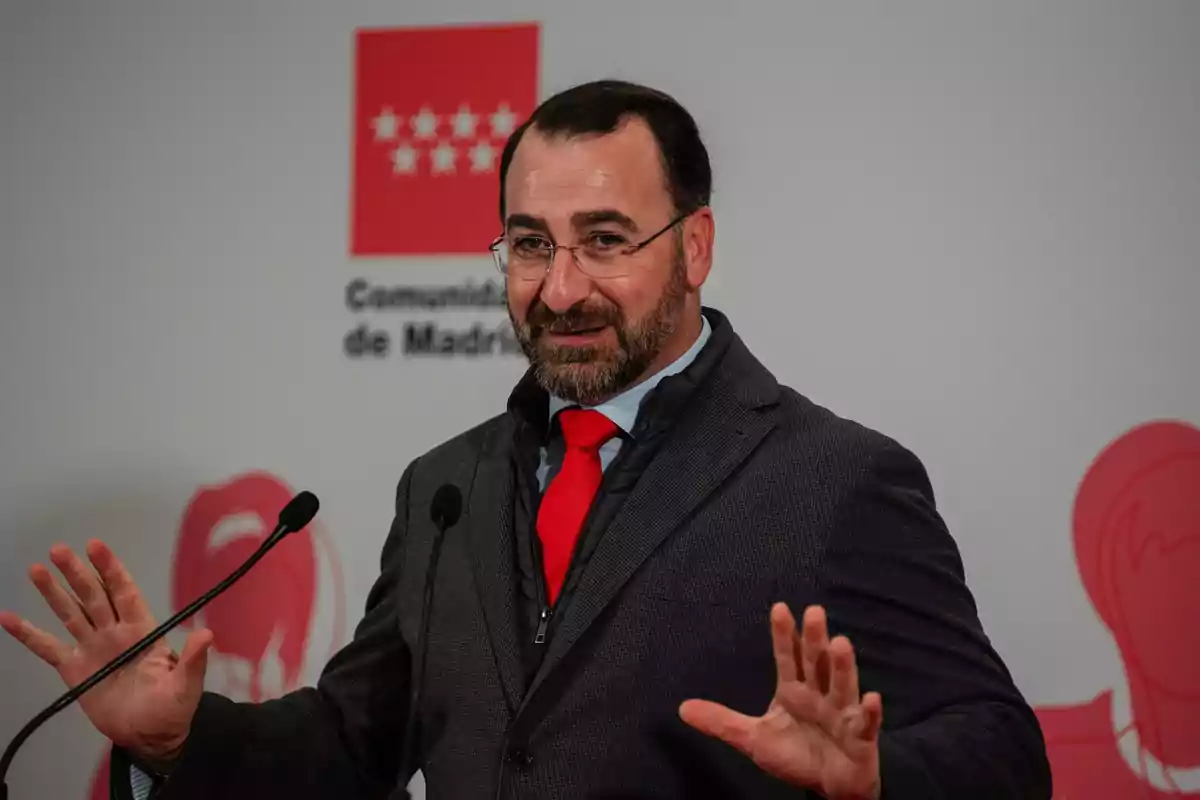 Hombre con barba y gafas hablando en una rueda de prensa con fondo de la Comunidad de Madrid