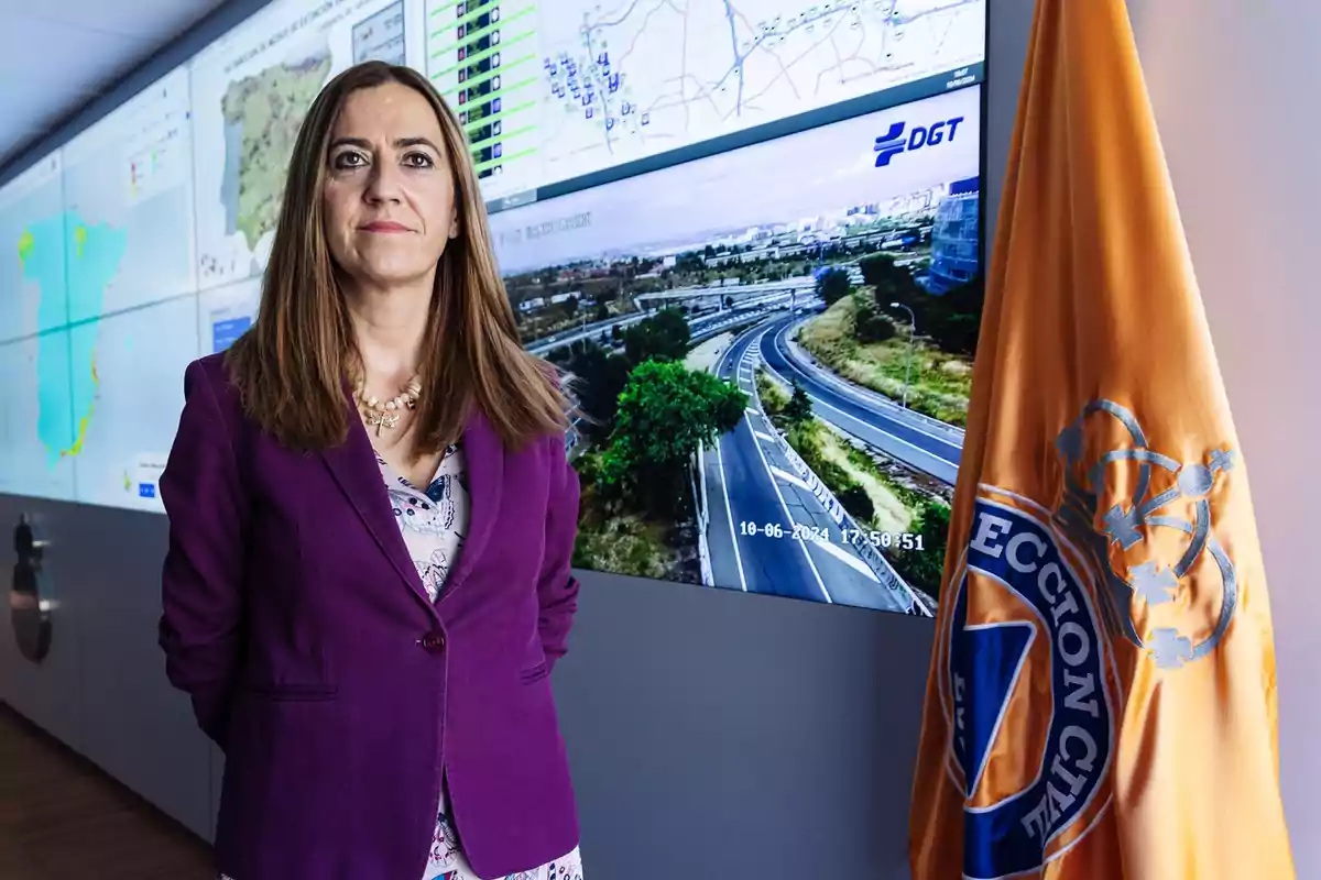 Mujer con chaqueta morada de pie junto a una bandera naranja en una sala de control con pantallas que muestran mapas y una carretera Mujer con chaqueta morada de pie junto a una bandera naranja en una sala de control con pantallas que muestran mapas y una carretera