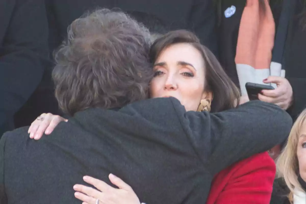 Dos personas se abrazan con fuerza en un ambiente solemne mientras una mujer observa al frente con expresión seria Dos personas se abrazan con fuerza en un ambiente solemne mientras una mujer observa al frente con expresión seria