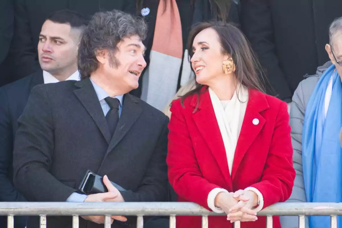 Un hombre con saco oscuro y una mujer con abrigo rojo conversan sonrientes en un evento al aire libre rodeados de otras personas. Un hombre con saco oscuro y una mujer con abrigo rojo conversan sonrientes en un evento al aire libre rodeados de otras personas.