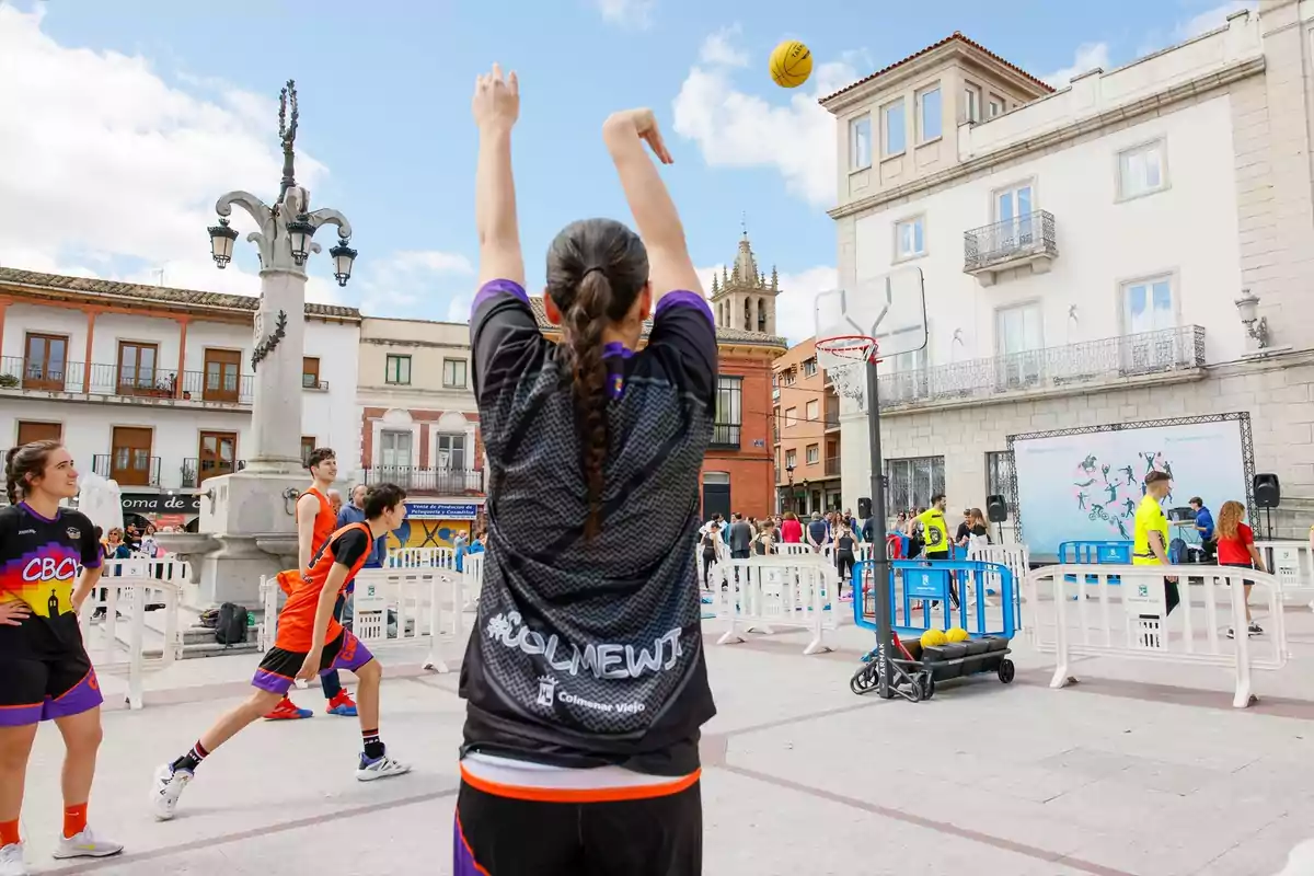 Una joven lanza un balón de baloncesto hacia la canasta en una cancha al aire libre situada en una plaza rodeada de edificios y personas observando el partido