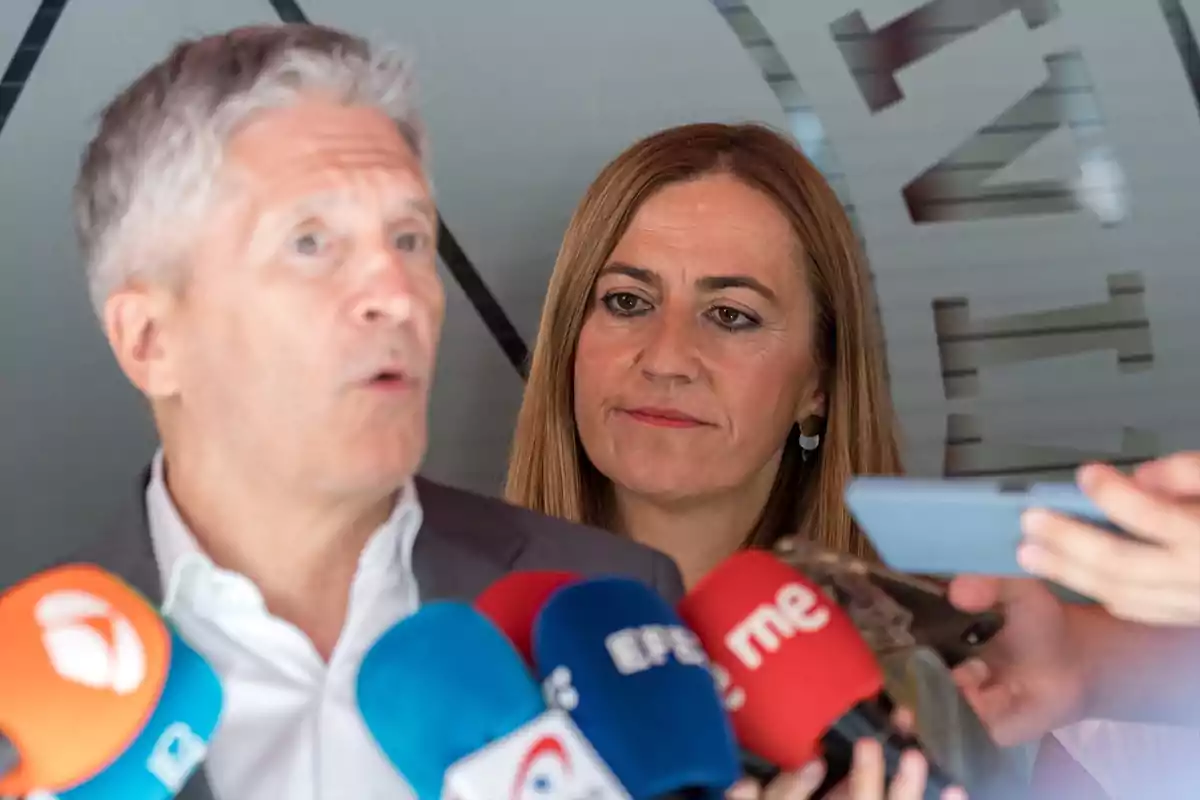 Un hombre hablando frente a varios micrófonos de diferentes medios de comunicación mientras una mujer lo observa atentamente en segundo plano