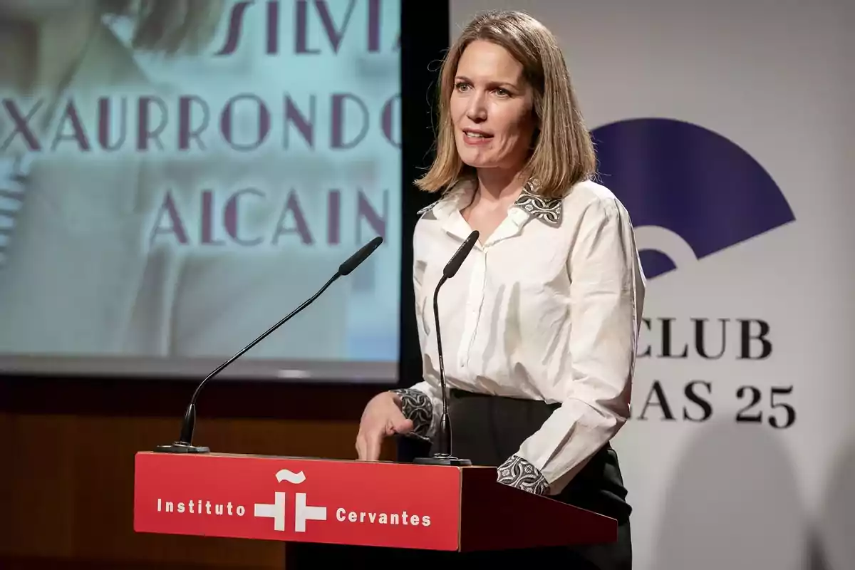 Mujer de cabello rubio y camisa blanca hablando en un atril del Instituto Cervantes durante un evento o conferencia