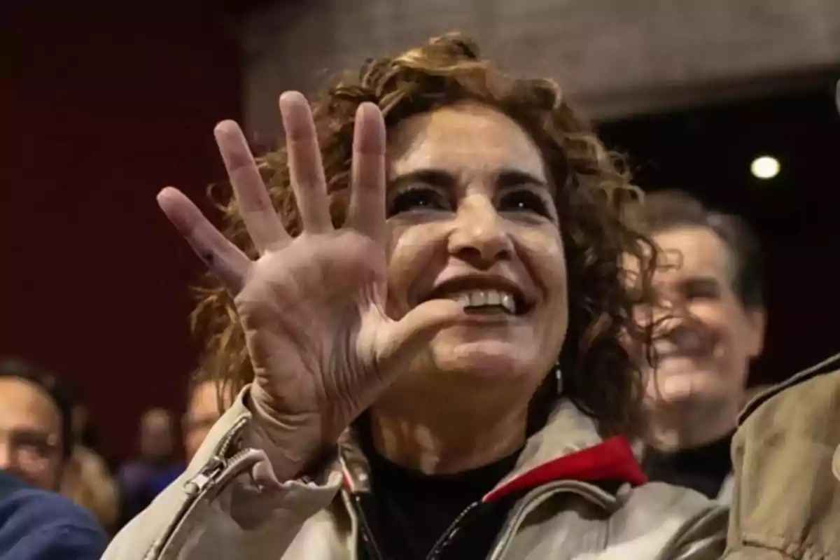 Mujer sonriente con cabello rizado levantando la mano y mostrando cinco dedos en un ambiente interior con personas al fondo Mujer sonriente con cabello rizado levantando la mano y mostrando cinco dedos en un ambiente interior con personas al fondo