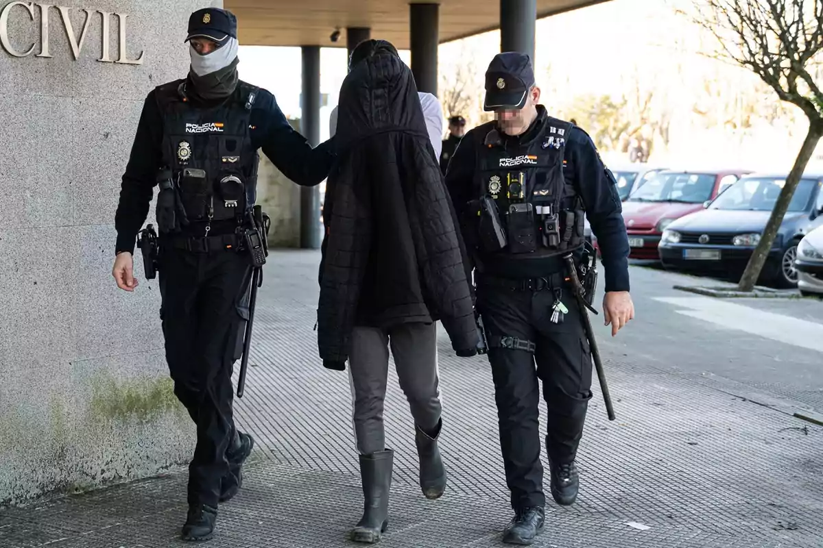 Dos policías escoltan a una persona detenida que lleva la cabeza cubierta con una chaqueta mientras caminan por la acera junto a un edificio y varios autos estacionados.