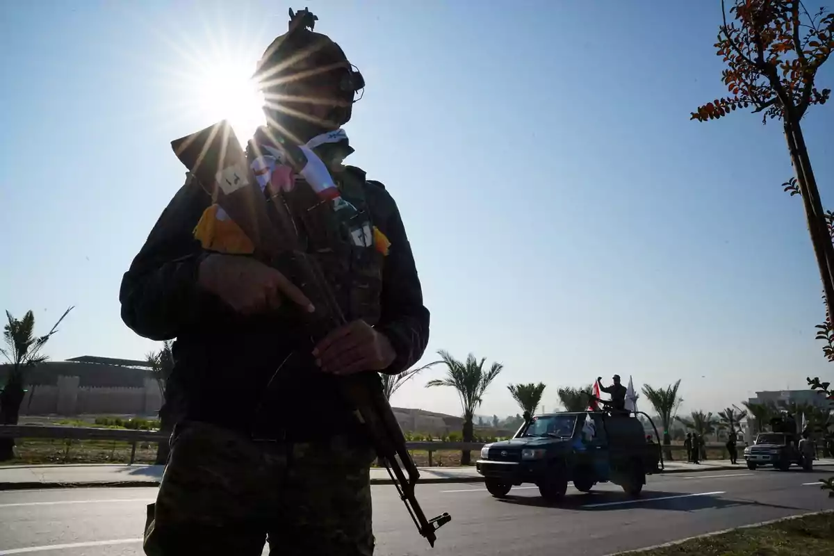 Un soldado armado de pie en una calle con el sol brillando detrás de él y vehículos militares circulando al fondo Un soldado armado de pie en una calle con el sol brillando detrás de él y vehículos militares circulando al fondo