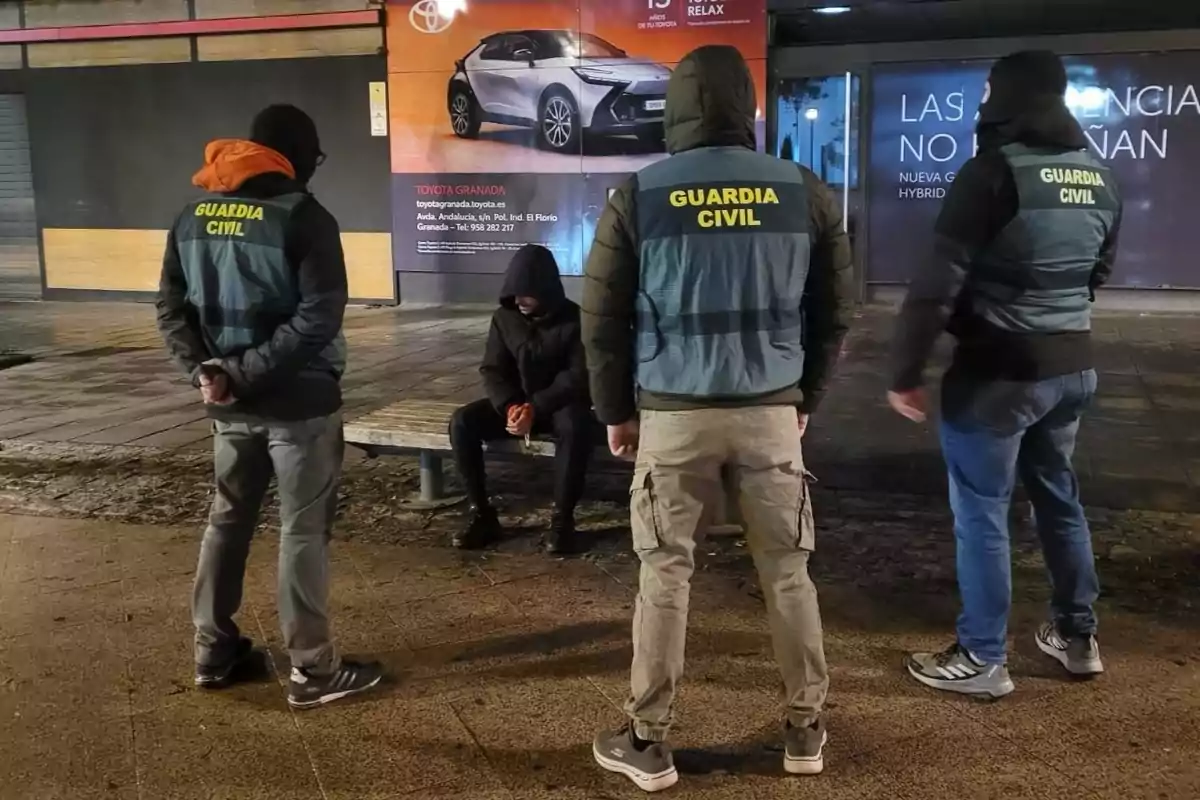 Tres agentes de la Guardia Civil rodean a una persona sentada en una banca en la calle durante la noche Tres agentes de la Guardia Civil rodean a una persona sentada en una banca en la calle durante la noche