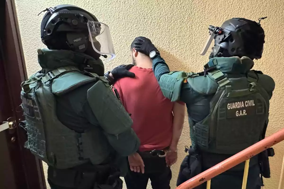 Dos agentes de la Guardia Civil con equipo táctico detienen a un hombre esposado en un pasillo junto a una pared amarilla.