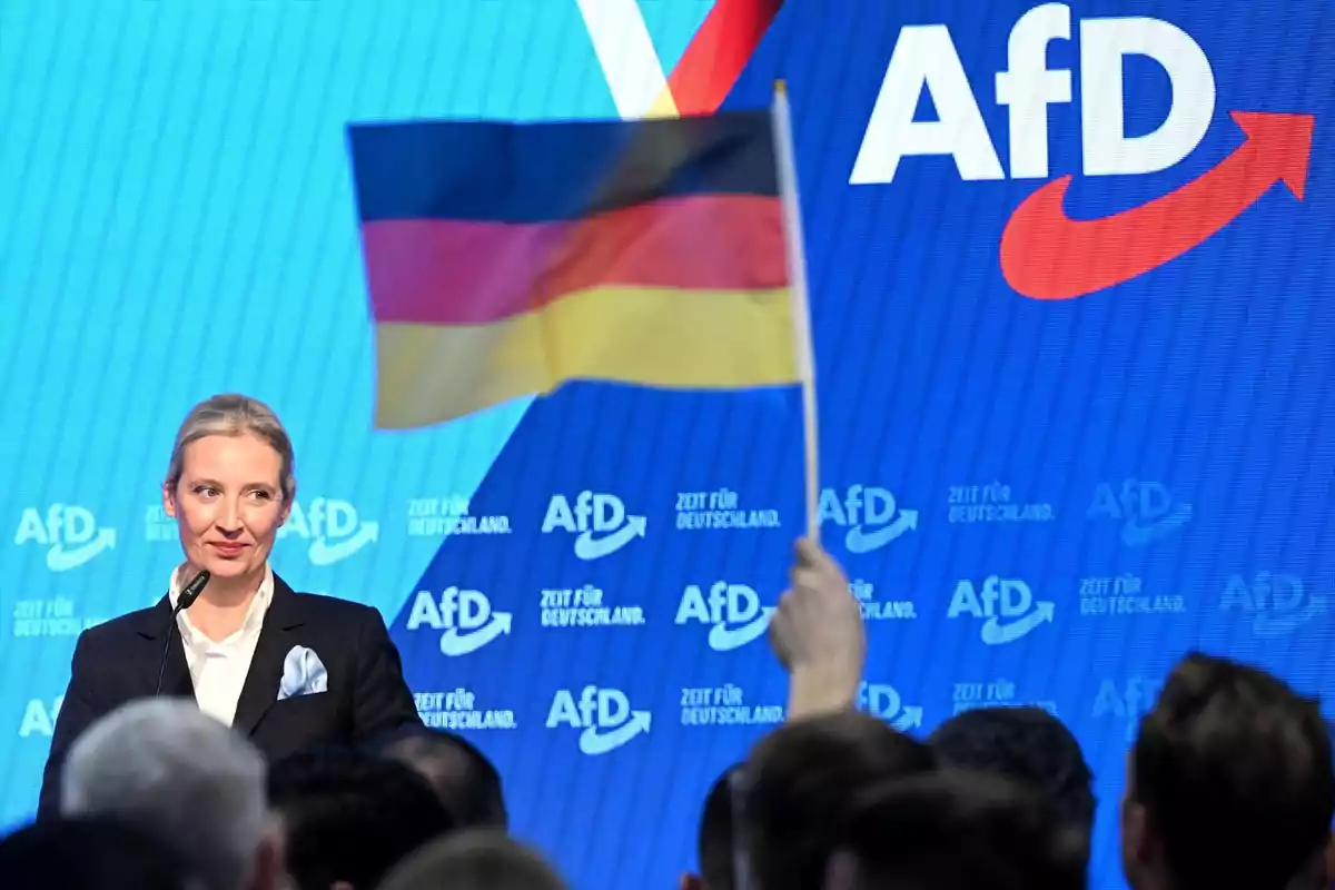 Una mujer de traje oscuro habla en un podio frente a una pantalla azul con el logo de AfD mientras una persona en el público sostiene una bandera de Alemania
