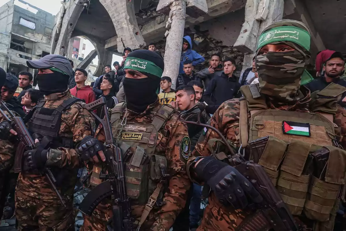 Tres hombres armados con uniforme militar y rostros cubiertos están de pie frente a un grupo de personas y edificios destruidos Tres hombres armados con uniforme militar y rostros cubiertos están de pie frente a un grupo de personas y edificios destruidos