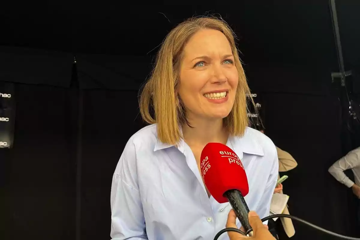 Mujer sonriente con camisa azul claro siendo entrevistada con un micrófono rojo de Europa Press frente a un fondo oscuro Mujer sonriente con camisa azul claro siendo entrevistada con un micrófono rojo de Europa Press frente a un fondo oscuro
