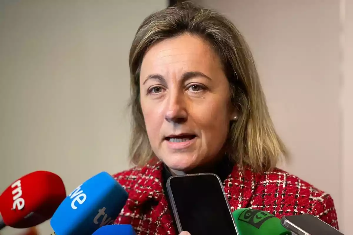 Mujer de cabello rubio oscuro y chaqueta roja hablando frente a varios micrófonos de diferentes medios de comunicación Mujer de cabello rubio oscuro y chaqueta roja hablando frente a varios micrófonos de diferentes medios de comunicación