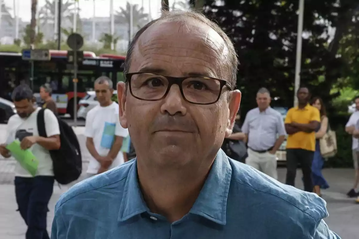 Hombre de mediana edad con gafas y camisa azul mirando a la cámara en una calle con varias personas y un autobús al fondo Hombre de mediana edad con gafas y camisa azul mirando a la cámara en una calle con varias personas y un autobús al fondo