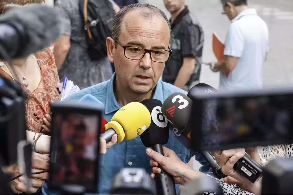 Un hombre con gafas y camisa azul responde preguntas de varios periodistas que sostienen micrófonos y grabadoras frente a él en la calle. Un hombre con gafas y camisa azul responde preguntas de varios periodistas que sostienen micrófonos y grabadoras frente a él en la calle.