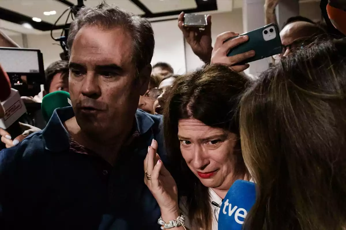 Una mujer con expresión de angustia es escoltada por un hombre mientras varias personas y periodistas con micrófonos y teléfonos móviles los rodean.