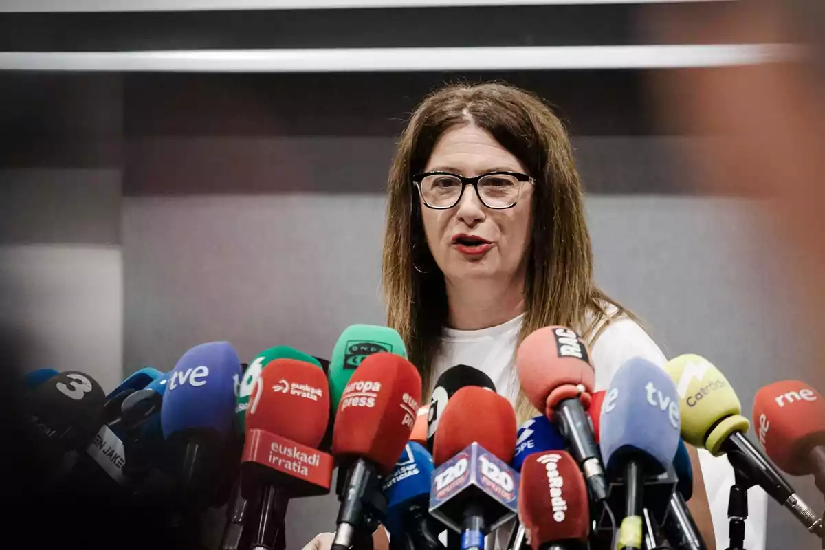 Mujer de cabello largo y gafas hablando frente a numerosos micrófonos de diferentes medios de comunicación en una rueda de prensa Mujer de cabello largo y gafas hablando frente a numerosos micrófonos de diferentes medios de comunicación en una rueda de prensa