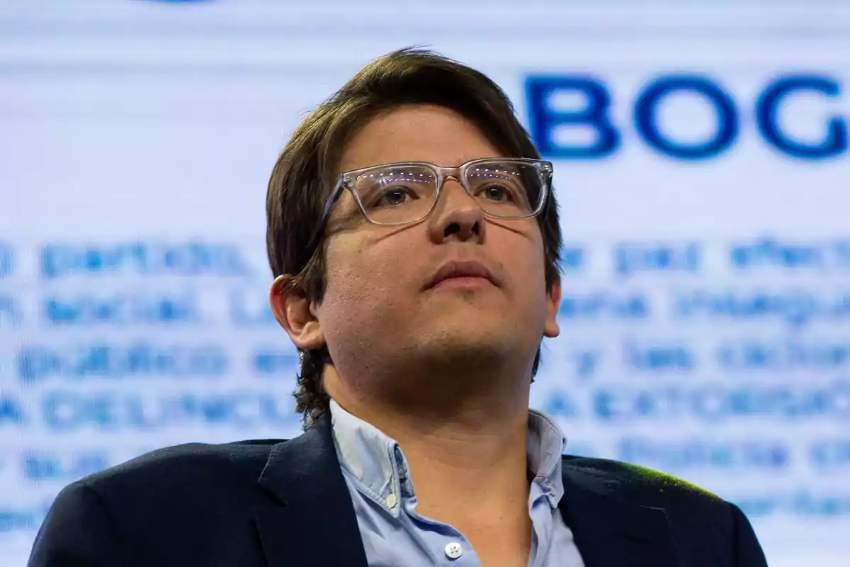 Hombre con gafas transparentes y cabello castaño mirando hacia arriba con expresión seria, fondo desenfocado con letras azules grandes