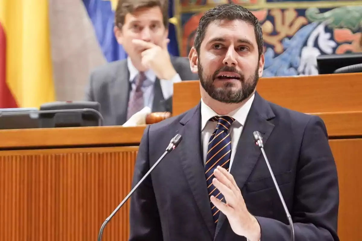 Hombre con barba y traje hablando en un podio con micrófonos en un entorno parlamentario mientras otra persona lo observa al fondo Hombre con barba y traje hablando en un podio con micrófonos en un entorno parlamentario mientras otra persona lo observa al fondo