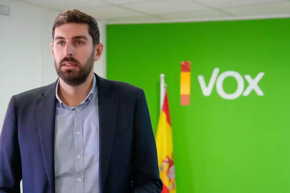 Hombre con barba y chaqueta oscura de pie frente a una pared verde con el logo de Vox y una bandera de España al fondo Hombre con barba y chaqueta oscura de pie frente a una pared verde con el logo de Vox y una bandera de España al fondo