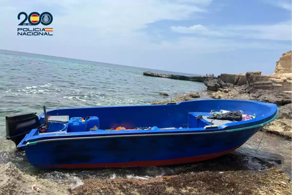 Una lancha azul con motor fuera de borda está varada en la orilla rocosa de una playa, con bidones y objetos en su interior, bajo un cielo parcialmente nublado y el mar de fondo, en la esquina superior izquierda aparece el logo de la Policía Nacional de España.