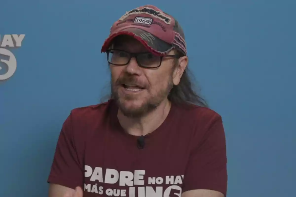 Hombre con gafas y gorra roja desgastada, lleva una camiseta burdeos con letras blancas y está frente a un fondo azul Hombre con gafas y gorra roja desgastada, lleva una camiseta burdeos con letras blancas y está frente a un fondo azul