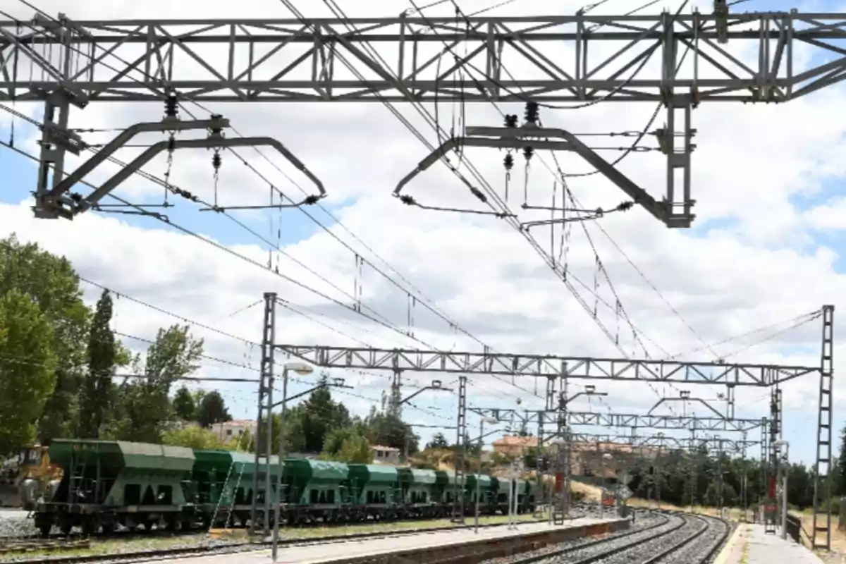 Vagones de tren de carga verdes estacionados en una vía férrea electrificada bajo una estructura de cables y postes metálicos Vagones de tren de carga verdes estacionados en una vía férrea electrificada bajo una estructura de cables y postes metálicos
