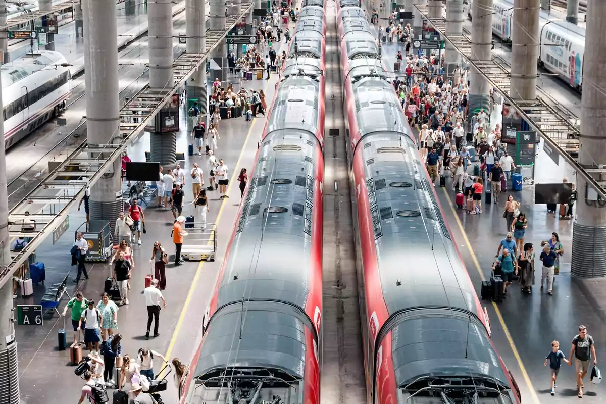 Andén de estación de tren con dos trenes rojos y muchas personas caminando y esperando con maletas