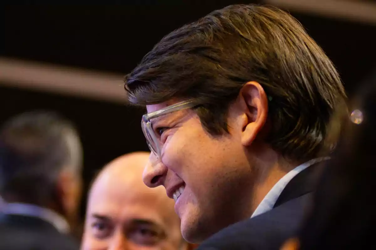 Hombre joven con gafas transparentes sonriendo en un ambiente interior junto a otras personas Hombre joven con gafas transparentes sonriendo en un ambiente interior junto a otras personas