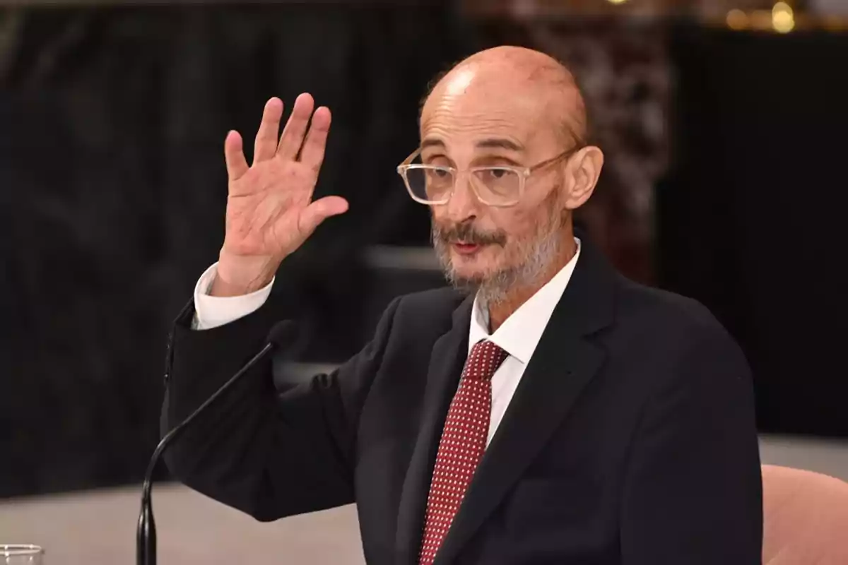Hombre calvo con gafas y barba canosa usando traje oscuro y corbata roja, levantando la mano derecha mientras habla frente a un micrófono.
