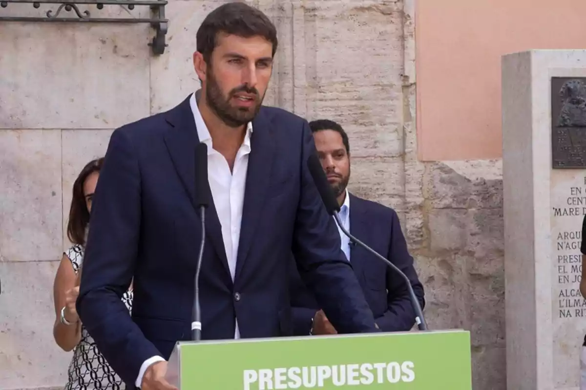 Un hombre con barba y traje azul habla en un atril verde con la palabra presupuestos mientras otras personas lo observan en un entorno exterior de piedra