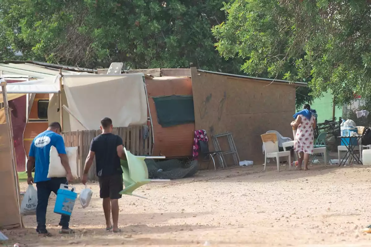 Varias personas se encuentran en un campamento improvisado con casas hechas de materiales sencillos y muebles al aire libre rodeados de árboles