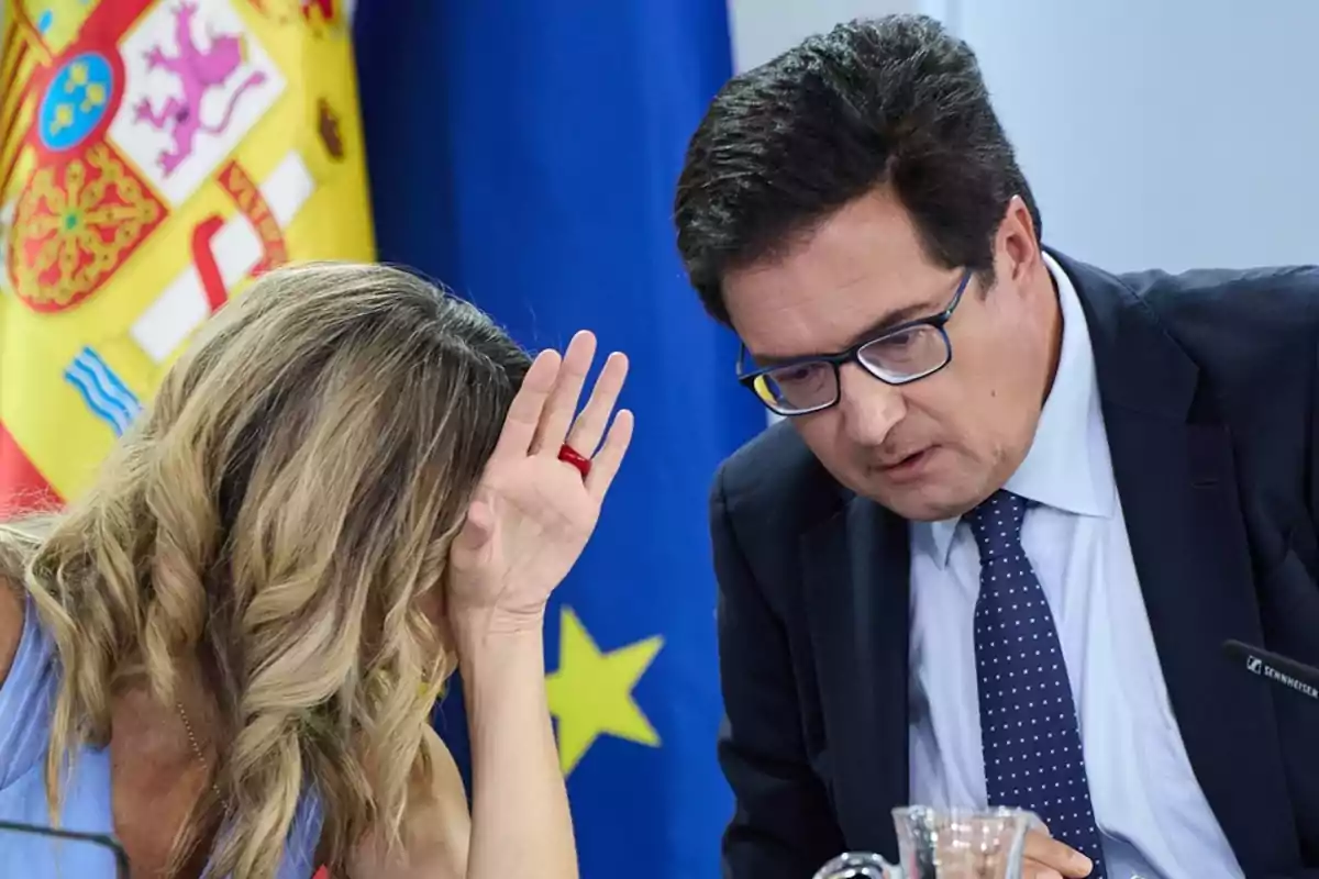 Dos personas conversan en una reunión formal frente a las banderas de España y la Unión Europea, una de ellas se cubre parcialmente el rostro con la mano. Dos personas conversan en una reunión formal frente a las banderas de España y la Unión Europea, una de ellas se cubre parcialmente el rostro con la mano.