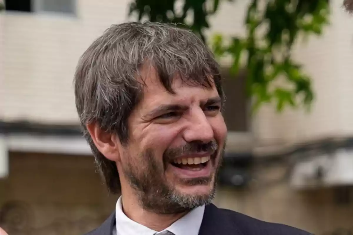 Hombre de cabello canoso y barba sonríe al aire libre con fondo de árboles y edificio desenfocado