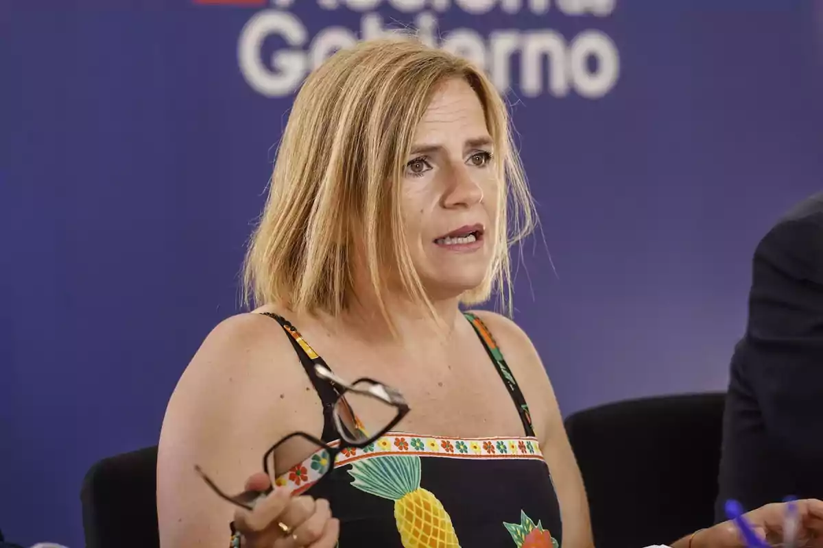 Mujer rubia con vestido de piña sosteniendo unos lentes mientras habla frente a un fondo azul con letras blancas desenfocadas