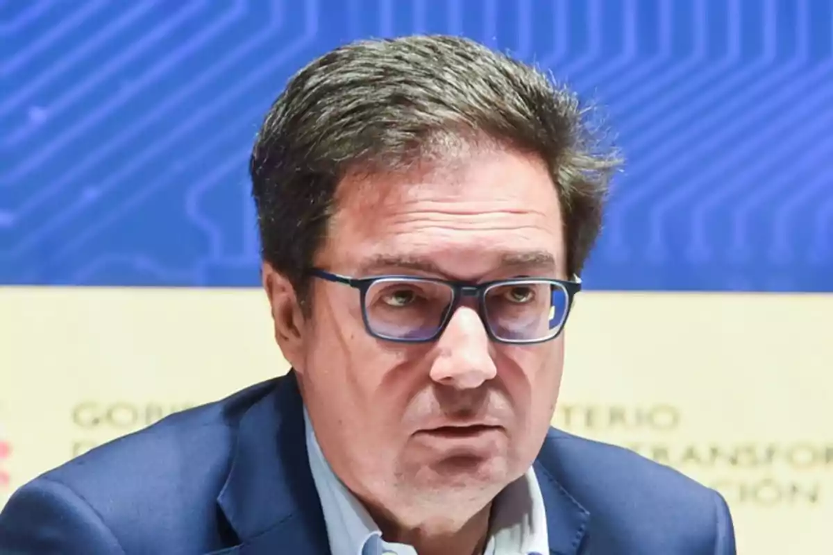 Hombre de cabello corto y gafas oscuras con expresión seria en un entorno profesional con fondo azul y amarillo Hombre de cabello corto y gafas oscuras con expresión seria en un entorno profesional con fondo azul y amarillo