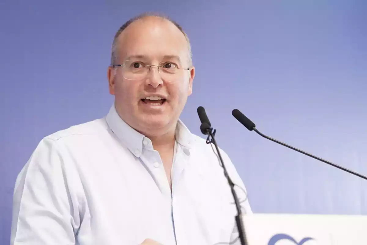 Hombre con gafas y camisa clara hablando frente a dos micrófonos sobre un fondo azul
