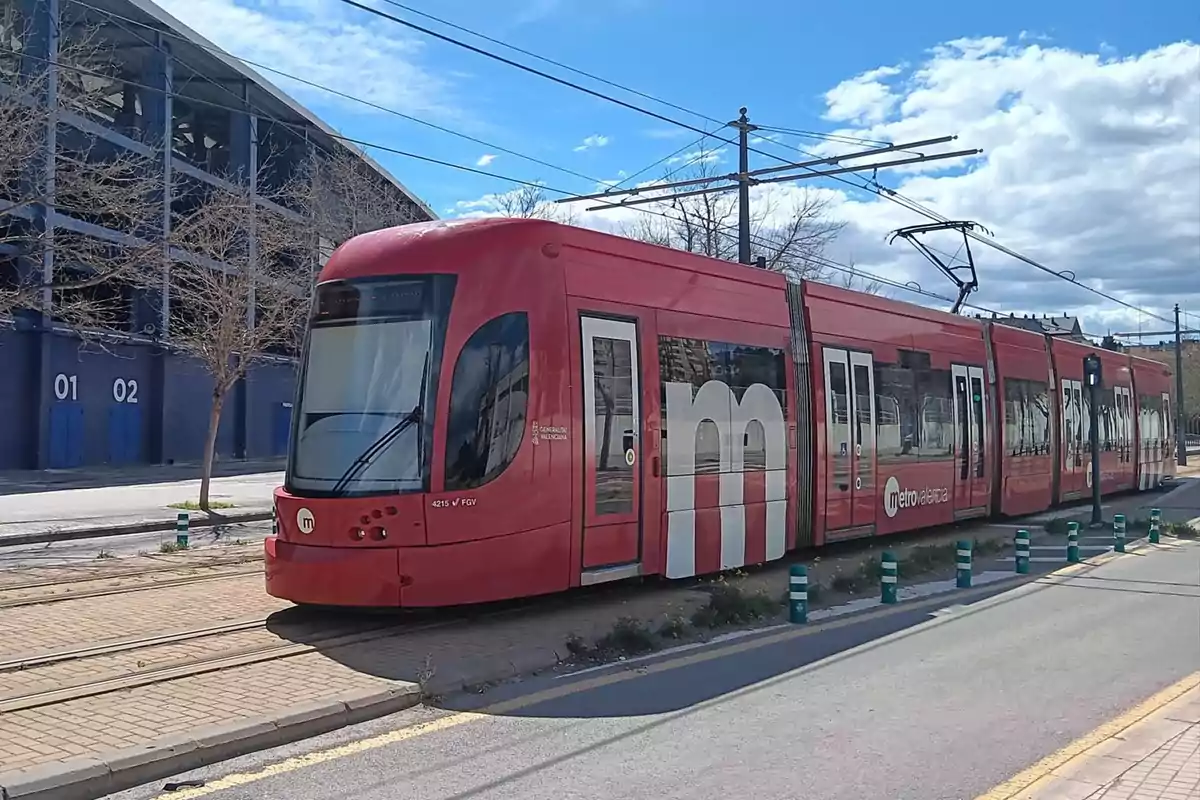 Tranvía rojo de Metrovalencia circulando por una vía junto a un edificio azul en un día soleado con algunas nubes en el cielo Tranvía rojo de Metrovalencia circulando por una vía junto a un edificio azul en un día soleado con algunas nubes en el cielo