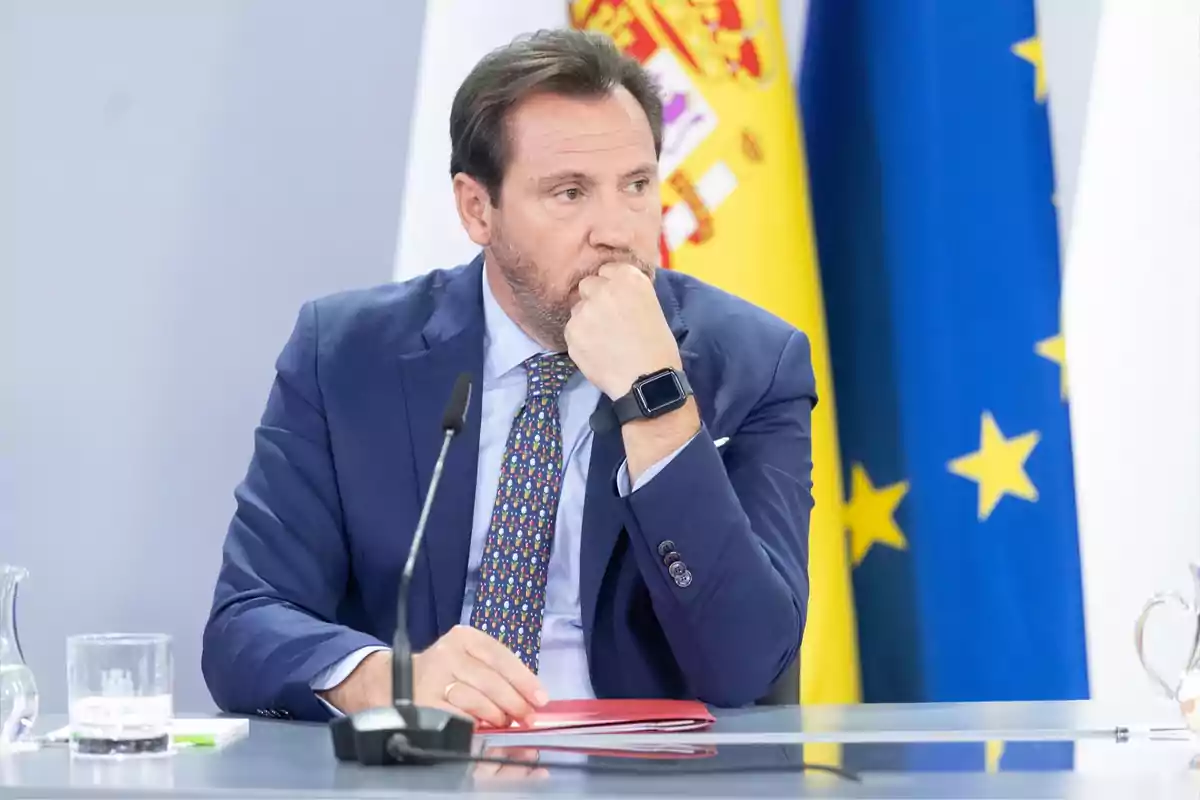 Hombre con traje azul sentado en una mesa de conferencia con banderas de España y la Unión Europea al fondo