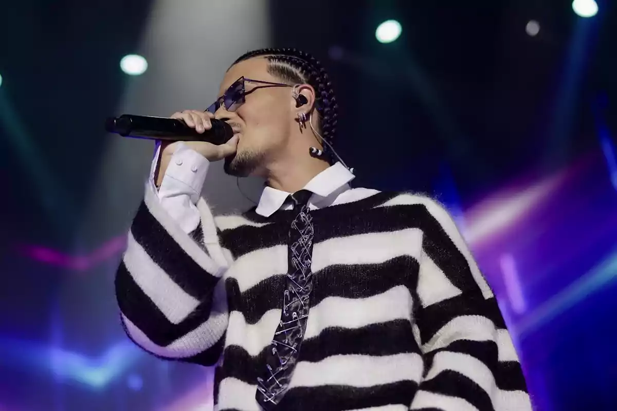 Cantante con gafas y trenzas actuando en el escenario con un suéter de rayas blancas y negras y corbata decorada con seguros, luces de colores al fondo