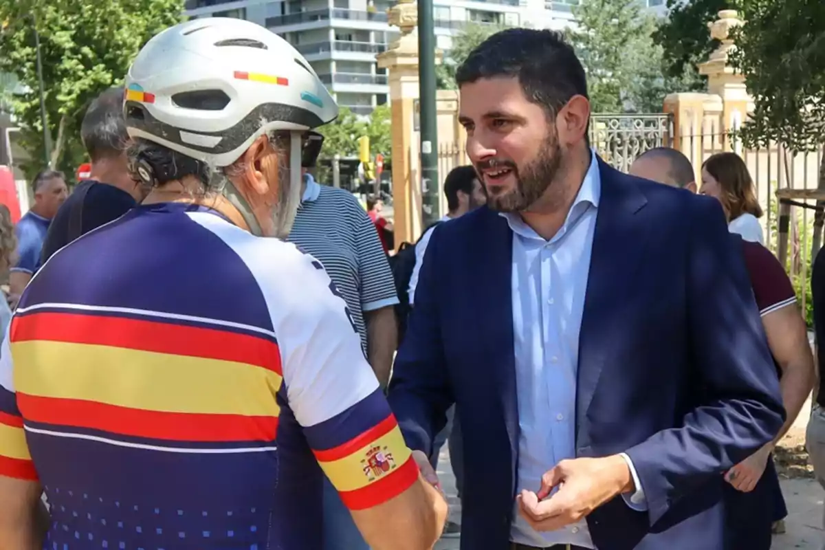 Un hombre con casco y ropa de ciclista saluda a otro hombre con barba y traje azul en un entorno urbano con varias personas al fondo