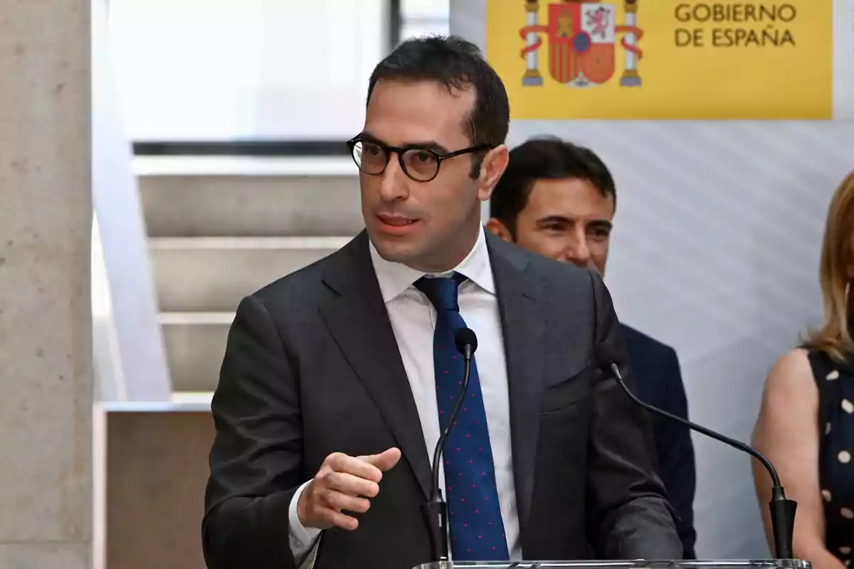Hombre con gafas y traje hablando en un podio durante un evento oficial del Gobierno de España Hombre con gafas y traje hablando en un podio durante un evento oficial del Gobierno de España
