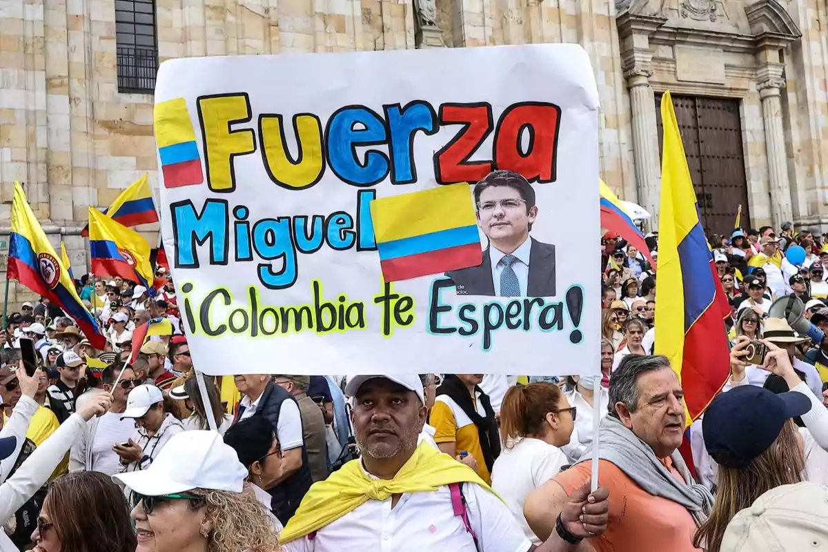 Manifestación en la que una persona sostiene un cartel colorido con el mensaje Fuerza Miguel Colombia te espera acompañado de una foto de un hombre y una bandera de Colombia, rodeado de una multitud con banderas colombianas frente a un edificio antiguo Manifestación en la que una persona sostiene un cartel colorido con el mensaje Fuerza Miguel Colombia te espera acompañado de una foto de un hombre y una bandera de Colombia, rodeado de una multitud con banderas colombianas frente a un edificio antiguo
