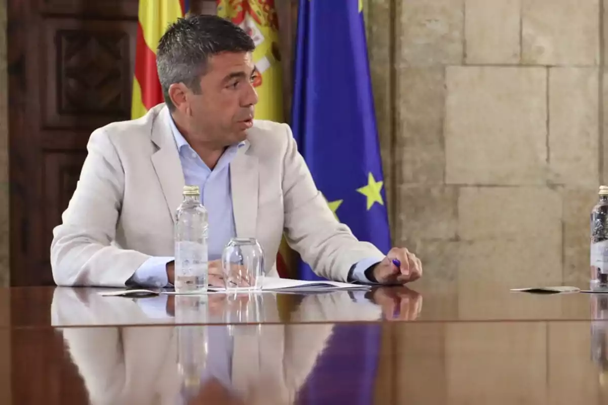 Hombre de cabello corto y canoso con chaqueta clara sentado en una mesa de reuniones con banderas de España y la Unión Europea al fondo