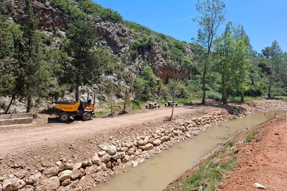 Camino de tierra junto a un canal de agua con una máquina de construcción estacionada y mesas de picnic rodeadas de árboles y montañas. Camino de tierra junto a un canal de agua con una máquina de construcción estacionada y mesas de picnic rodeadas de árboles y montañas.