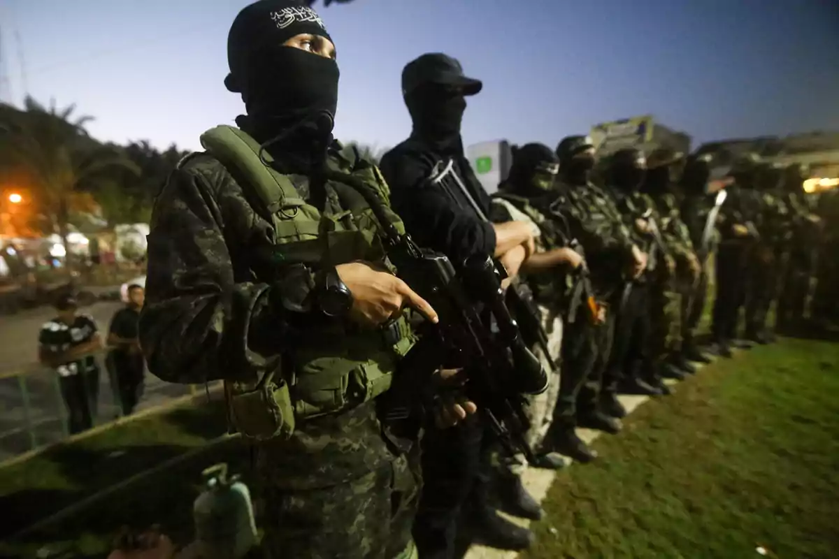 Grupo de hombres armados con uniformes militares y pasamontañas alineados al aire libre durante el atardecer Grupo de hombres armados con uniformes militares y pasamontañas alineados al aire libre durante el atardecer