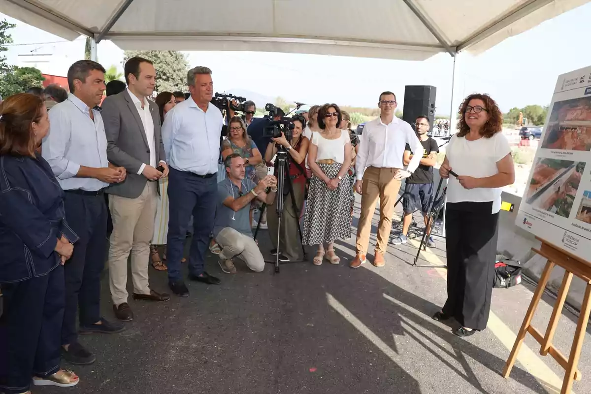 Un grupo de personas asiste a una presentación al aire libre bajo una carpa, mientras una mujer explica información mostrada en un panel con imágenes de obras públicas.