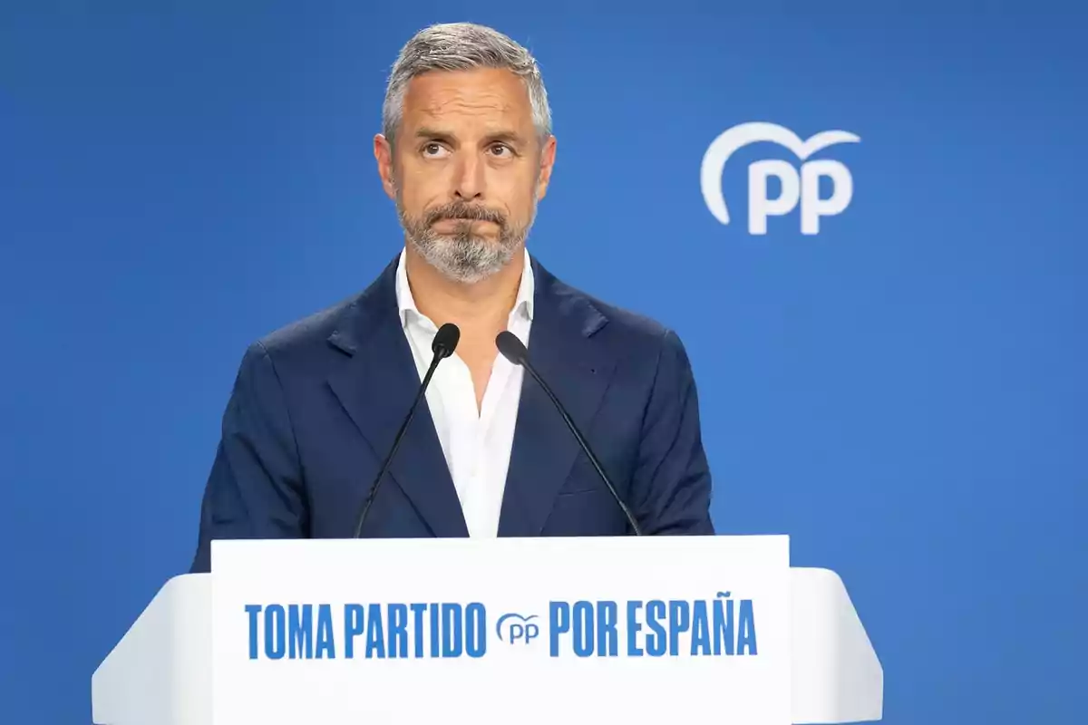 Hombre de cabello canoso y barba hablando en un atril con el texto Toma partido por España y fondo azul con el logo del Partido Popular Hombre de cabello canoso y barba hablando en un atril con el texto Toma partido por España y fondo azul con el logo del Partido Popular