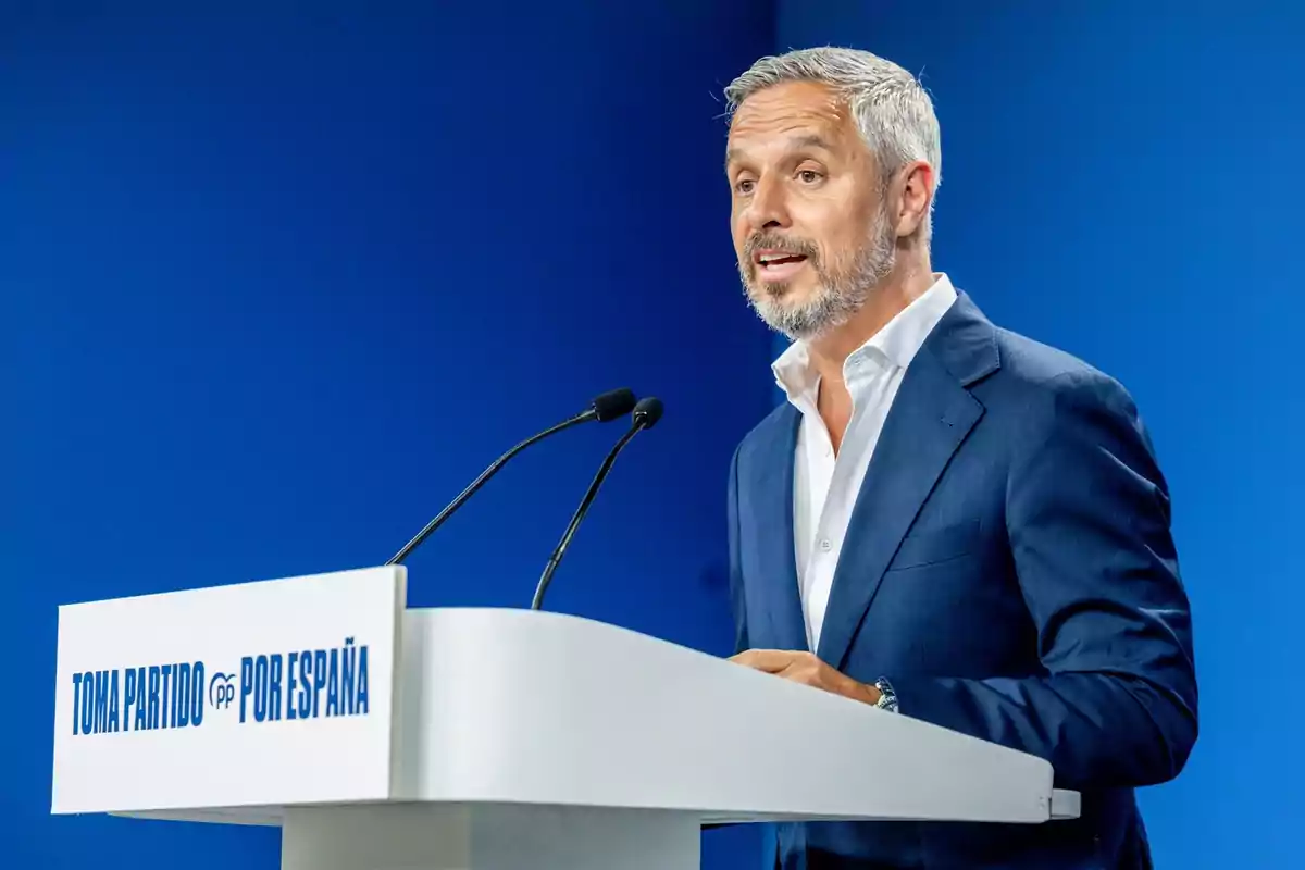 Hombre de cabello canoso y barba hablando en un atril blanco con el texto Toma Partido por España sobre un fondo azul